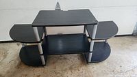 Front view of black laminated particle board entertainment stand with three shelves and plastic legs on concrete floor.