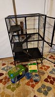 Full view of large metal wire ferret/rat cage showing door open, platforms, and interior damage. Accessories on floor beside cage including small travel cage, exercise balls, and wood shavings bag.