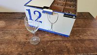 Opened box with one wine glass goblet displayed outside in front of the box showing the style and size.