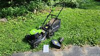 Full view of Greenworks electric corded push mower on grass with grass collection bag attached and side chute placed next to it, along with a manual.