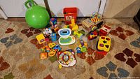 Various baby and toddler toys arranged on carpet floor including bouncing ball, educational laptop toy, toy train and shape sorter.