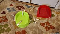 Photo showing a red freestanding squishy rubber booster seat and a green Safety 1st strap-on booster seat with attached white straps, placed on a carpeted floor.