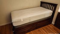 Twin size bed frame in dark wood finish with visible scratches, two pull-out drawers open and closed, mattress on top with white cover without mattress protector.