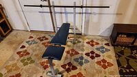 Full view of adjustable weight bench press with bar on a patterned rug in a garage or storage space.