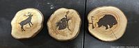 Three wooden coasters laid out horizontally, each a round wood slice with bark edges and natural rings. Left coaster shows a deer silhouette, middle a pineapple sketch, and right a rhinoceros silhouette.