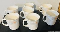 Six white ceramic CorningWare coffee cups with grey band and orange/beige floral design arranged in two rows.