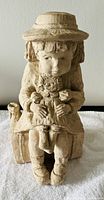 Beige textured sculpture of a seated girl wearing a hat holding a doll, against neutral background.