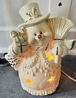 White ceramic snowman with light-up star cutouts, holding a broom and wearing a hat with holly decoration.