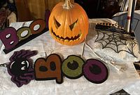 Photo of Halloween decorations on a white cloth including a lighted orange ceramic pumpkin with carved face, foam glitter signs spelling 'BOO' and 'BOOO', a purple spider glitter sign, and a packaged spider web sparkle sign.