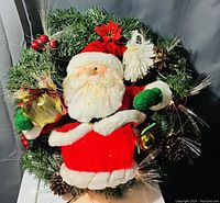 Front view of the Santa Claus Christmas wreath showing plush Santa holding a gold bag with holiday greenery and decorations.