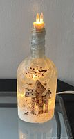 Glass bottle lamp with light inside showing painted winter scene with houses and snowman, topped with twine and twin candle-shaped lights.