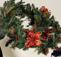 Close-up photo showing green artificial pine garland decorated with pinecones, red berries, and glittery red poinsettia flowers with integrated string lights.