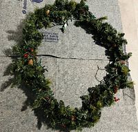 Green Christmas garland laid in a circular shape, decorated with pine cones, red berries, and lights.
