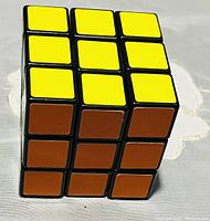 Front angled photo showing the 3x3 Rubik's Cube with yellow stickers on top and brown stickers on the front side, placed on a white fabric surface.