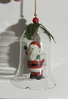 Close-up photo of a clear glass bell ornament containing a Santa Claus figurine, hanging against a white wall.