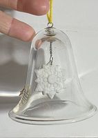 Clear glass bell ornament with white snowflake inside shown held by fingers for scale. The ornament is hanging from a yellow loop.