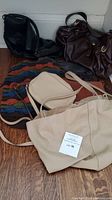 Set of five handbags including black woven leather bag, beige large tote, brown handbag, multicolor woven bag, and small beige crossbody bag on floor.