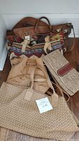 Five handbags arranged on floor including a leather satchel, tapestry bag, woven tote, bucket bag, and straw clutch