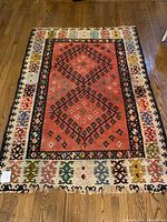 Full view of the rectangular rug laid flat on wooden floor showing central coral-red field with black geometric medallions and colorful outer border.