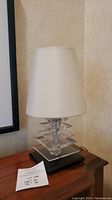 Front view of the lamp showing white tapered fabric shade over layered acrylic body on black base, with lot tag visible.