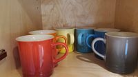 Side angle view of six Le Creuset stoneware mugs in assorted colors arranged in a wooden shelf.