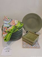 Stack of 12 green crackle side plates with 6 colorful patterned napkins on top, 2 square green dishes stacked, and patterned trivet beneath dishes.