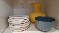 Stack of 8 white ceramic plates, yellow vase and blue bowl shown side by side.
