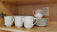 Shelf with 6 white coffee mugs, 9 saucers stacked, and 1 white creamer pitcher