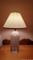 Front view of the lamp showing white ceramic base and pleated fabric shade with lamp illuminated
