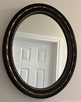 Oval wall mirror showing the black frame with gold details, reflecting a white door and part of a wall.
