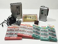 Two vintage Panasonic handheld cassette and microcassette recorders with earphones, multiple unopened Maxell microcassette packs, and a cleaner and care kit arranged on white background.
