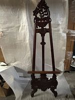 Front view of the tall mahogany wood easel with ornate carved top and base, showcasing the overall design and height.