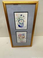 First image shows two floral watercolour paintings in a single gold frame under glass with blue-grey matting. The top painting depicts flowers in a blue vase, bottom painting colorful flowers in a white container.