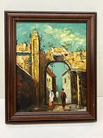 Framed oil painting showing a large stone archway with two figures walking through it, buildings on either side, blue sky with clouds above.