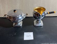 Front view of chrome creamer and lidded sugar bowl with Bakelite handles and square bases placed on black surface.