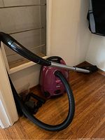 Purple Bissell Zing canister vacuum cleaner with attached hose and metal wand, shown standing on wooden floor near wall.
