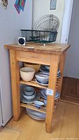 Butcher block table with shelves holding bowls and dishes (not included), wooden trivet and wire basket rack on top.