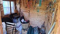 View inside shed showing hanging tools, storage bins, rolled mats, basket, and brooms leaning against wall