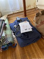 Folded navy blue electric blanket with an attached plug, controller, and instruction sheet on top of it, laying on a wooden floor next to woven rugs.