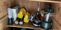 Photo showing variety of flashlights and camping lanterns arranged on wooden shelf including yellow handheld flashlights, metallic small flashlights, black flashlight, two camping lanterns, and a package of batteries.