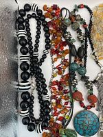 Multiple necklaces with various bead colors including black, white striped, multicolored glass beads, and turquoise stone pendant on black cord.