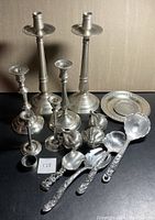 Multiple pewter serving items on a dark surface showing candlesticks, salt and pepper shakers, plate, and serving utensils