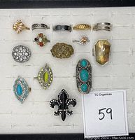 Top view of 14 assorted rings arranged in a display tray showing various colors, shapes, and stone types including turquoise, clear rhinestones, amber-colored stone, black enamel, green and pink stones.