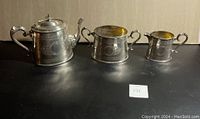 Full view of the three-piece Russian silver plated tea set - teapot, sugar bowl and creamer with ornate handles and detailed engraving on dark surface.