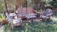 Wide view showing one wooden lawn bench, two wooden chairs, and three side tables set on grassy yard against a wooden fence.