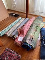 Five rolled throw rugs on floor near window, showing colors and fringe ends.