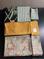 Tablecloths and napkins laid flat showing patterns and colors. Includes green checkered, yellow floral, and blue-gray vintage floral patterns.