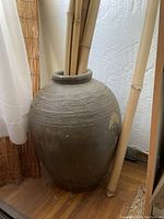 Tall decorative vase about 3 feet tall, sitting on wooden floor next to window with white curtain, holding tall bamboo-like sticks. Vase is textured with horizontal ridges near the neck and shows natural wear and discoloration.