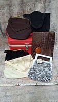 Seven vintage bags arranged on a red train case including brown clasp bag, textured black bag, brown clutch with crocodile texture, white beaded bag, black velvet bag, and white/navy bag with beads.