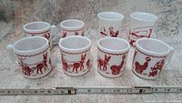 Front and side perspective of 8 vintage Hazel Atlas glass pieces, 2 tumblers and 6 mugs, with red cowboy and circus designs on white glass.